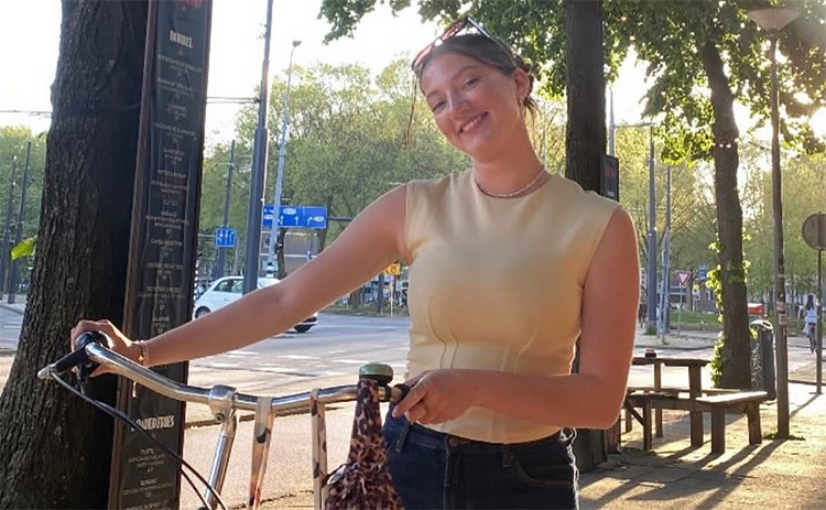 Olivia Farnworth holding a bicycle by the handlebars outdoors on a tree-lined street.