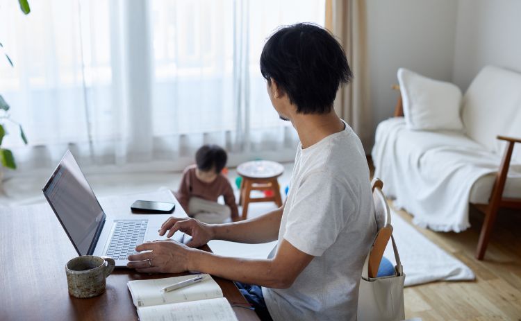 Adult working on a laptop at a dining table with notebooks and a mug, while a young child plays on the floor in the background of a bright living room.