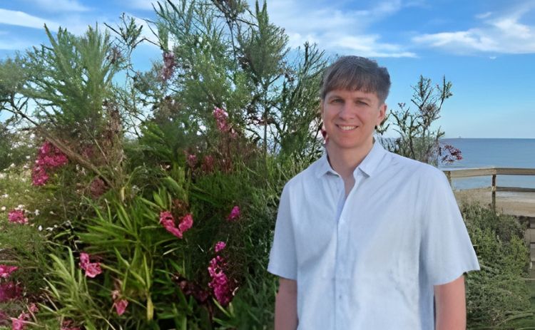NUBS student Sam Carr wearing a light short-sleeved shirt stands smiling beside flowering shrubs on a sunny coastal path, with the sea and blue sky in the background.
