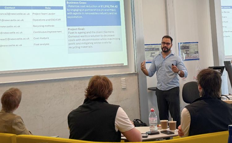  A person giving a presentation in a classroom, standing beside a projected slide while attendees sit at tables listening.