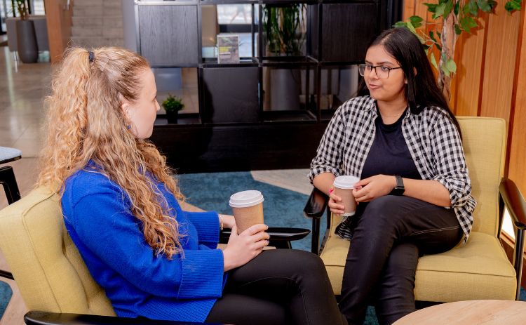Light-skinned student in a bright blue sweater and dark-skinned student in a black-and-white checkered shirt sit in a modern business school café with green armchairs, wooden walls, plants, shelves, and a blue rug, chatting over takeaway coffee cups.
