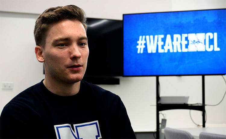 Tom Horvat wearing a dark T-shirt with the Newcastle University sports logo. A TV in the background shows text "#WEARENCL"