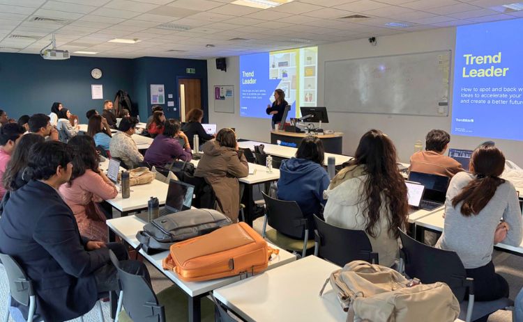 A diverse group of students sit at desks with laptops and notebooks in a bright classroom, listening to a presentation. At the front, a speaker stands near a large screen displaying a slide titled “Trend Leader” with text and graphics.