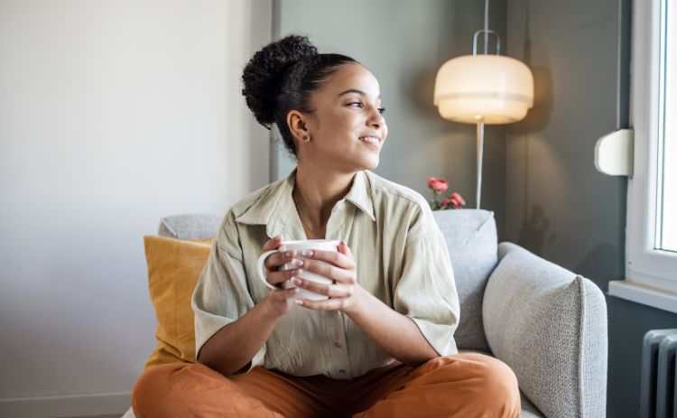 A dark-skinned woman sitting in a grey armchair, holding a white mug with both hands while looking out of a nearby window.