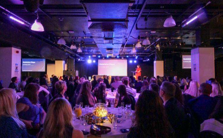 A wide-angle shot of the Workplace Awards 2025 ceremony. Attendees sit at round tables watching a speaker on stage beneath bright purple lighting. A presentation slide shows the name Madeleine Chiya from Go Volunteer NEXT.