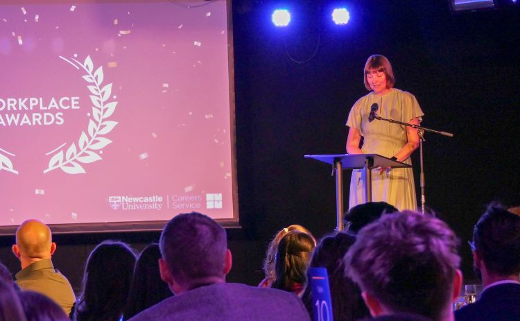 A light-skinned woman in a grey dress speaks at a podium on stage at the Workplace Awards 2025. The screen behind her displays the event logo and Newcastle University and Careers Service branding. Audience members watch from round tables in the foreground.