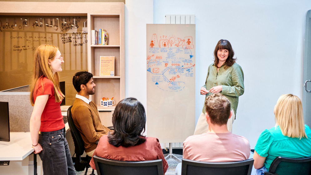A female stands in front of a whiteboard and other students presenting