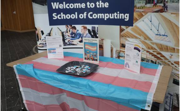 A table covered in a trans pride flag. On top are three signs with resources for trans students and staff and a bowl filled with keyrings with the trans pride flag