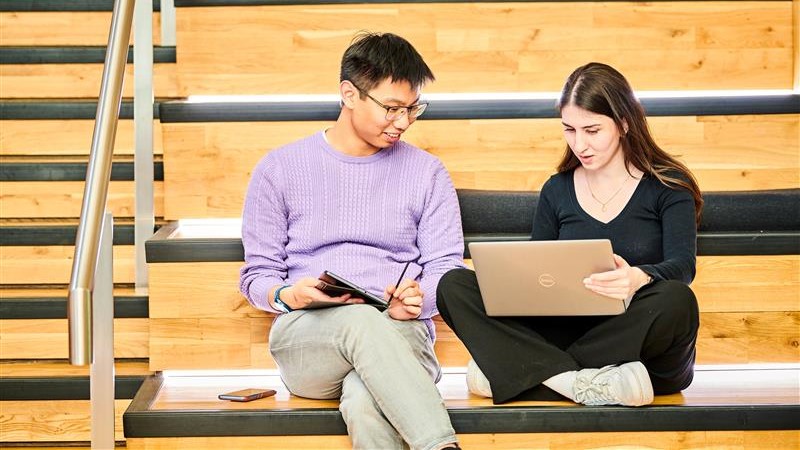 A man and a woman sit on a bench next to some stairs, working together on a laptop.