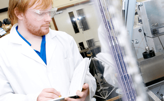 A man wearing goggles and a labcoat making notes in a lab.