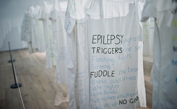 Rows of white clothing hanging in a gallery displaying quotes about epilepsy