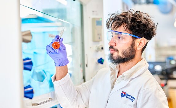 Person in a lab coat and safety goggles examining a vial with orange substance in a research lab with equipment and storage containers