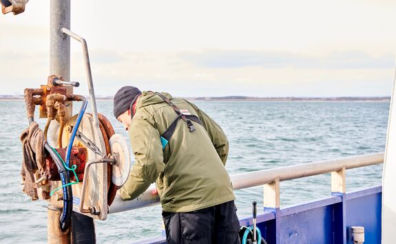 Person on a boat conducting acoustic signalling research, leaning over the railing with marine equipment, wearing a green jacket and black hat, with water and distant land in the background