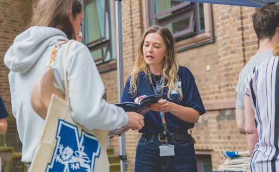 A university staff member in a navy shirt is speaking to a student and showing an information booklet at an outdoor information stall, with other people standing nearby against a background of a brick building.